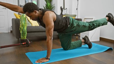 Afrika kökenli Amerikalı bir adam oturma odasında egzersiz yapıyor, mavi bir paspasın üzerinde yoga yapıyor, fitness ve denge gösterisi yapıyor..