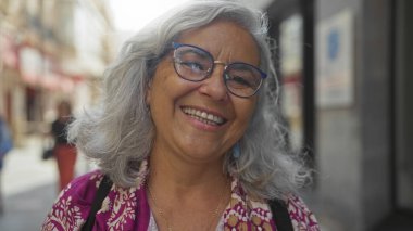 Gri saçlı ve gözlüklü yaşlı bir kadın, gün boyunca renkli giysiler içinde bir caddede gülümsüyor.