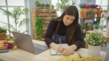 Hispanic woman takes notes in a flower shop with a laptop and vibrant flowers in the background. clipart