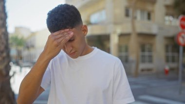 Güneşli bir şehir caddesinde sorunlu görünen genç bir adam, şehir stresi ve tefekkür hissi uyandırıyor..
