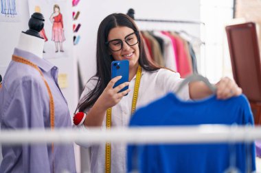 Young beautiful hispanic woman tailor make photo by smartphone to sweater at atelier