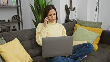 Evde çalışan İspanyol bir kadın rahat bir oturma odasında telefonla konuşurken dizüstü bilgisayar kullanıyor..