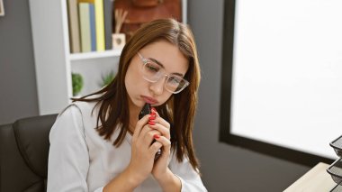 Ofiste, elinde gözlük ve beyaz bir bluz ile telefon tutan düşünceli genç bir kadın..