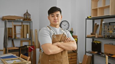 İyi organize edilmiş bir marangozluk atölyesinde kollarını kavuşturan önlüklü kendinden emin Asyalı genç bir adam..