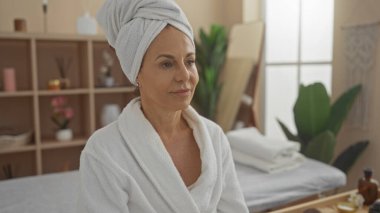 Havlu sarığı ve bornozu olan orta yaşlı bir kadın, iyi aydınlatılmış bir sağlık merkezinde sakin bir ifadeyle evde oturuyor.