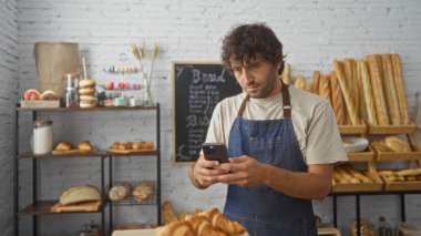 Fırındaki genç adam telefonunu kontrol ediyor. Etrafı ekmek ve hamur işleriyle çevrili.
