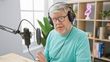 Kulaklık takan gri saçlı bir adam radyo stüdyosunda mikrofona konuşuyor. Profesyonelliği ve odaklanmayı işaret ediyor..