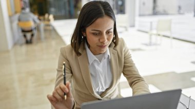 A focused hispanic woman in business attire works attentively on a laptop in a modern office environment. clipart