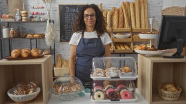 Fırında duran bir kadın gözlük ve önlükle çeşitli hamur işleri ve ekmeklerle dükkânın samimi ve hoş atmosferini sergiliyor..