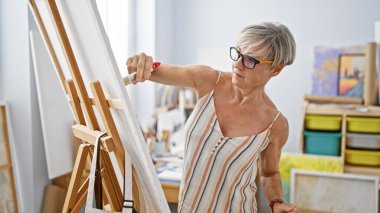 Parlak bir sanat stüdyosunda tuvale resim yapan odaklanmış olgun bir kadın sanatçı.