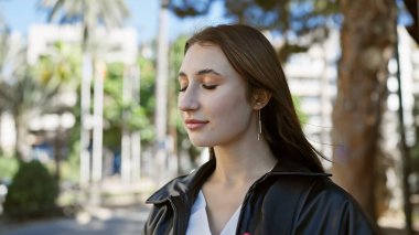 Düşünceli genç bir kadın güneşli bir şehir ortamında poz veriyor, sakin bakışları şehrin telaşının ortasında sükuneti yansıtıyor..