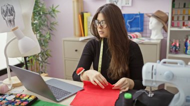 Canlı bir terzi dükkanında dizüstü bilgisayarı, dikiş makinesi ve renkli iplikleri olan İspanyol bir kadın..