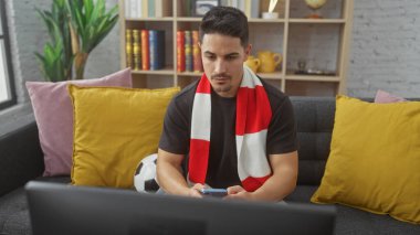 Oturma odasında futbol eşarbı ve laptopuyla akıllı telefon kullanan sakallı İspanyol bir adam..