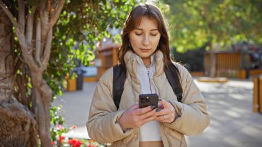 Güneşli bir şehir parkında akıllı telefon kullanan genç bir kadın. Etrafı yeşillik ve çiçeklerle çevrili..