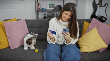 İspanyol kadın telefona kredi kartıyla gülümsüyor, köpek kanepenin yanında, modern oturma odasında..