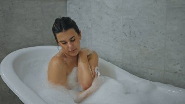 Genç yetişkin bir kadın, ev içi banyo ortamında köpük banyosunda dinleniyor ve sakinliğini ifade ediyor..