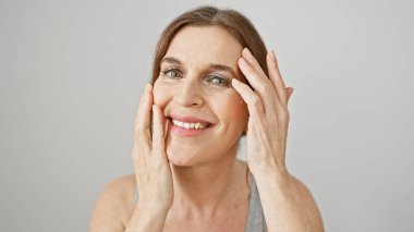 Neşeli, orta yaşlı bir kadın yüzüne dokunuyor, beyaz bir arkaplanda poz veriyor ve gülümsüyor..