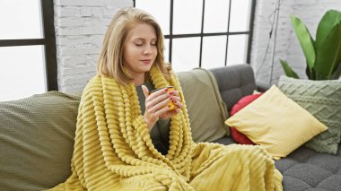 Sarışın, rahat bir battaniyeye sarılmış genç bir kadın, gri bir kanepede sıcak bir içeceğin tadını çıkarıyor..