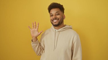 Neşeli Afro-Amerikan adam, günlük bej kazak giyip, parlak sarı bir arka plana selam veriyor..