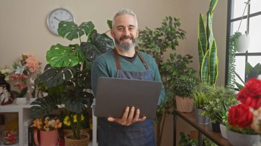 Sakallı adam çiçek dükkanında dizüstü bilgisayar tutuyor. Etrafı yeşil bitkilerle çevrili.