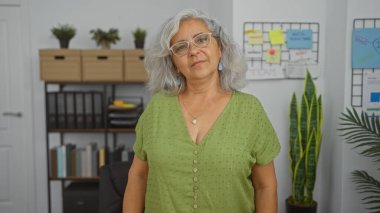 Yeşil bluzlu, gri saçlı ve gözlüklü yaşlı bir kadın, arka planda bitki, kitap rafları ve beyaz bir tahtayla organize bir ofis ortamında duruyor..