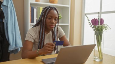 Odaklanmış örgü ören Afrikalı bir kadın rahat bir evde online ödeme için kredi kartı kullanıyor..