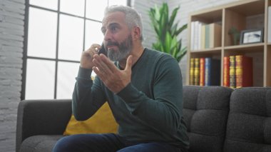 Olgun sakallı bir adam, rahat, modern bir oturma odasında telefonla konuşurken hareketli bir şekilde hareket eder..