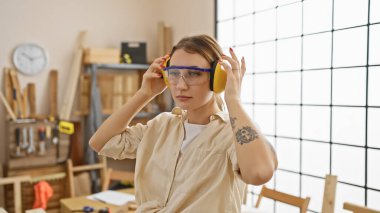 İyi aydınlatılmış bir marangozluk atölyesinde güvenlik gözlüğü ve kulaklık takan genç bir kadın.