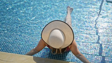 Genç bir kadın, güneşli bir yüzme havuzunun kenarında, hasır bir şapka takarak, boş zamanları ve yaz havasını simgeleyen bir şekilde rahatlıyor..