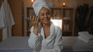 Sessiz bir spa odasında dinlenirken telefonundan sesli mesaj dinleyen bir kadın, kapalı bir ortamda mumlar ve yumuşak ışıklandırmayla rahatlık ve rahatlık gösteriyor..