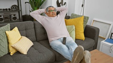 Renkli yastıkları ve ev dekorasyonu olan rahat bir oturma odasında gri koltukta dinlenen olgun bir adam.