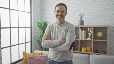 Gülümseyen orta yaşlı bir adam modern bir oturma odasında minimalist bir dekorla kollarını kavuşturmuş duruyor..