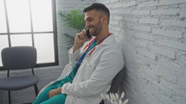 Sakallı, yakışıklı, genç, İspanyol bir doktor hastane bekleme odasında oturuyor. Gülümserken telefonla konuşuyor. Üzerinde beyaz bir ceket ve steteskop var..