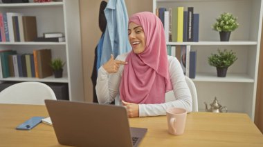 Sıcak bir ofis ortamında video görüşmesinde tesettürlü gülümseyen kadın kendini işaret ediyor.