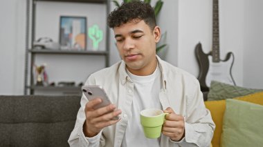 Modern oturma odasında akıllı telefon kullanırken sakallı yakışıklı bir genç kahve fincanı tutuyor.