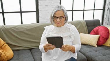 Gözlüklü yaşlı bir kadın rahat bir oturma odasında kanepede dinlenirken tablet kullanmaktan hoşlanır..