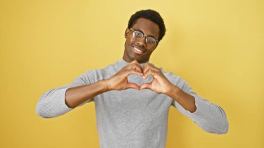 A smiling young african american man making a heart sign against a yellow isolated background. clipart