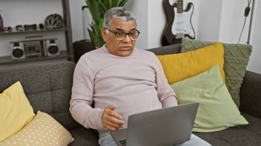 Gözlüklü, rahat bir oturma odasındaki koltukta dizüstü bilgisayar kullanan, yastıklı ve gitarlı olgun bir adam..