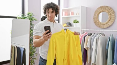 Beyaz polo giyen İspanyol bir adam elinde akıllı bir telefonla parlak bir elbise odasında sarı bir kapüşonluyu muayene ediyor..