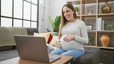 A smiling pregnant hispanic woman sits comfortably at home with a laptop, exuding maternal warmth in a cozy living room. clipart