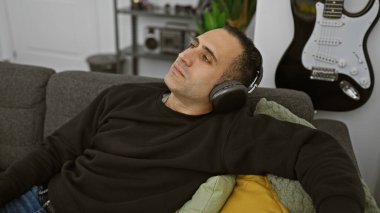 Rahatlamış İspanyol bir adam, rahat bir oturma odasında, bir gitar ve bir kanepenin yer aldığı kulaklıkla müzik dinlemekten hoşlanır..