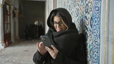 Siyah şallı, gülümseyen kadın, İstanbul 'daki Top Kapi Sarayı' nda süslü fayansların yanında akıllı telefon kullanıyor.
