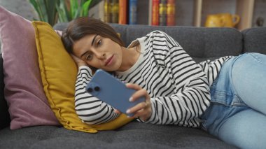 Güzel, genç bir İspanyol kadın renkli yastıklarla çevriliyken akıllı telefon kullanarak evinde uzanıyor..