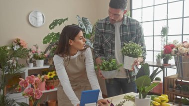 Bir kadın ve bir erkek çiçekçi dükkanında birlikte çalışıyorlar. Etrafı bitkilerle çevrili. Perakende bir ortamda iç mekan takım çalışmasını örnek alıyorlar..