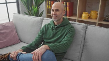 Sakallı, yeşil kapüşonlu, rahat bir oturma odasındaki gri kanepede oturmuş gülümseyen İspanyol kel adam..