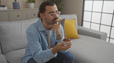 Oturma odasında oturan bıyıklı genç adam elinde bir kek ve açık mavi bir ceket ve gözlük takmış bir koltuğun önünde düşünüyor..