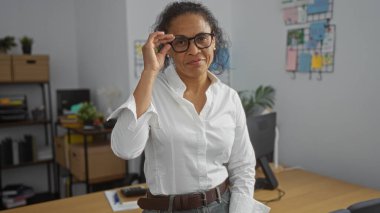 Ofiste gözlük takan bir kadın, Latin Amerikalı bir kadın, çalışma alanında olgun bir profesyonel, beyaz gömlek giyiyor, gülümsüyor, arka planda organize masalar kuruyor, etrafta canlı bitkiler var..