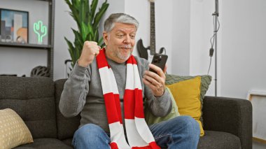 Gri saçlı neşeli orta yaşlı bir adam modern bir oturma odasında akıllı telefonuna bakarken kutlama yapıyor..
