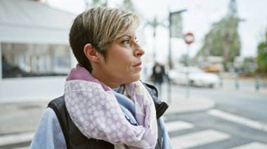 Kısa sarı saçlı, düşünceli bir İspanyol kadın şehir sokaklarında dikiliyor ve şehir hayatı hissi yayıyor..