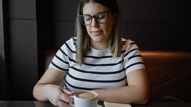 Düşünceli esmer bir kadın, elinde kahve fincanıyla bir kafede oturuyor..